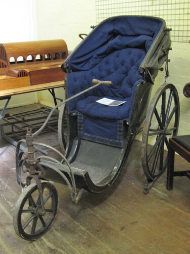 Bath chair in the Roman Baths museum store: St John's Museum Store, Upper Bristol Road, Bath. The three wheeled chair's seat is covered in blue fabric, with a black wooden exterior and a folding hood to protect the occupant against the elements.